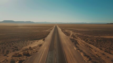 Fototapeta premium A straight, endless road cuts through a vast desert landscape under a clear, blue sky, creating a sense of isolation and adventure