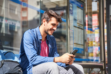 Bearded young man in denim shirt sitting at bus stop looking at his phone while waiting for bus in the background of modern city