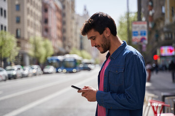Happy handsome brunette adult man using smartphone while standing beside traffic lane in daylight. Waiting for taxi cab order by application
