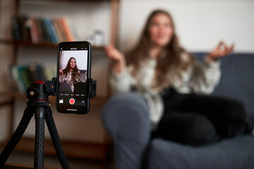 Selective focus on phone on tripod recording vlogger sitting on couch gesticulating and communicating with her followers, livestreaming for her blog in social media. Blogging as business