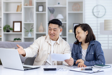 Young smiling Asian family, man and woman sitting at home at table in front of laptop and doing financial budget and documents.