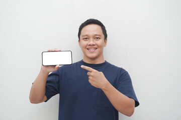 Young Asian man smiling and pointing to smartphone screen isolated on white background.  lifestyle and advertising concept. Men using blue shirt holding phone mockup