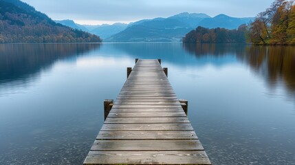 Fototapeta premium A wooden dock stretches across a still lake, leading towards mountains shrouded in mist