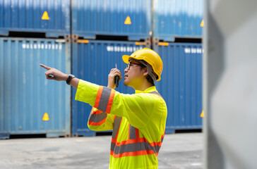 Asian worker in yellow safety helmet and reflective vast using walkie talkie in cargo shipping yard for communication container inspection. Man standing in front of blue shipping containers and pointi