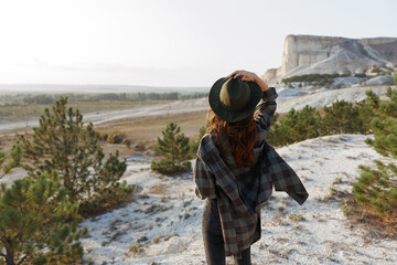 Woman in plaid shirt and hat stands on hill overlooking majestic mountain scenery