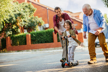 Father and grandfather teaching young boy to ride a scooter on suburban street