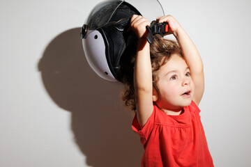 Cute brunette curly toddler girl in red t-shirt in white bike motorcycle helmet taking it off from...