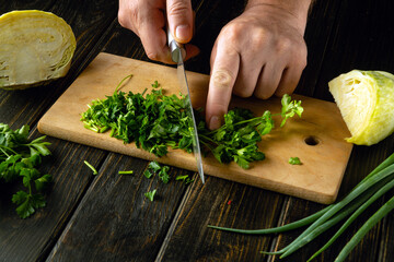 The cook hands use a knife to cut green parsley on a cutting board to prepare a vitamin salad....