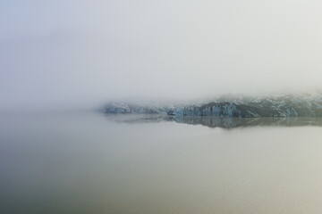 Naklejka premium A serene view of Sólheimajokull glacier in Iceland, shrouded in mist and reflecting in the calm glacial lagoon.