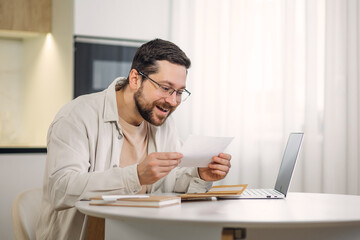Smiling young man holding paper bill letter while working at table with portable laptop indoors. Skilled caucasian freelancer feeling shock while reading received information at home interior.