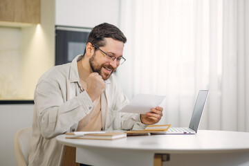 Successful caucasian male receive letter feeling happy to read positive news at home workplace. Bearded man in glasses expressing happy emotions on background of kitchen.