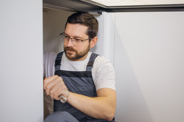 Fototapeta premium Calm caucasian plumber in grey coveralls securely attaching pipe details with screwdriver for long term service. Concerned repairman working with equipment indoors and installing sink parts.