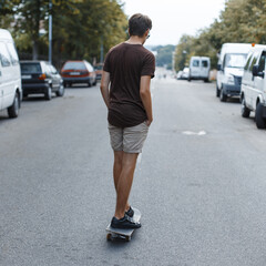 Young fashionable man in summer clothes with shorts and tank top riding a skateboard on the street
