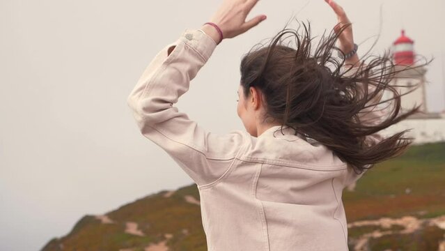 young beautiful woman tourist stands on the western edge of europe, cape cabo da roca in portugal. cloudy windy day. girl photographs the ocean by smartphone. hair flies and flutters
