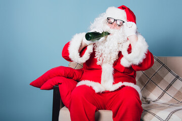 Funny fat Santa Claus watches TV on the couch at home and drinks beer. Blue background.