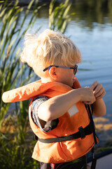 A young boy with blond hair and glasses puts on an orange life jacket by the water, surrounded by...