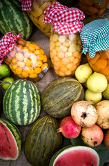Colorful array of fresh and jarred fruits, including watermelons, pears, pomegranates, and melons, displayed on burlap. Jars have checkered cloth lids, adding a rustic feel.