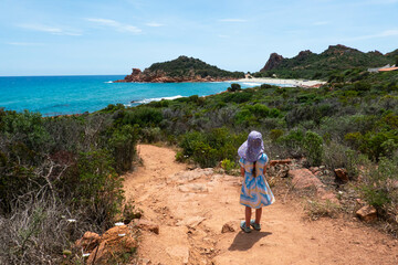 Little girl in dress standing on the path to wild hidden Su Sirboni beach and red rock formations in province of Nuoro on Sardinia (Sardegna) island in Italy. Tyrrhenian Sea.