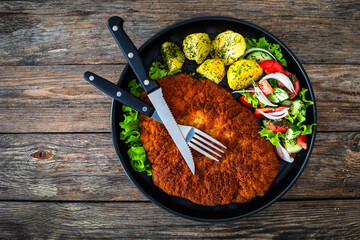 Crispy breaded fried pork chop, boiled potatoes and fresh vegetables served on black plate on wooden table
