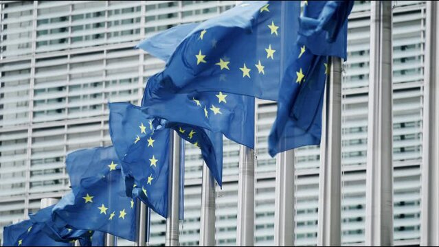 In Brussels, the political heart of the European Union, several EU flags flutter in harmony beside a contemporary glass building, embodying unity and progressiveness in a symbolism rich scene