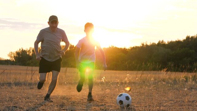kid boy running playing football with dad. kid kicking soccer ball run field. father chasing kid. Family football game at sunset