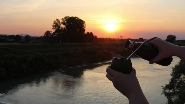Hands pouring hot water over yerba mate in a gourd at sunset, with a river landscape in the background