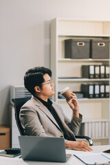 Naklejka premium Asian businessmen wearing glasses Sipping coffee while sitting on the computer screen. Concept is to sit and work smart Happy Bright and Relaxed atmosphere. Background is a document rack.
