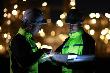 Two white man Electrical Engineers Discussing a laptop, light screen plan, and  Digital Tablet While Standing At a Dark Power Station During the Night