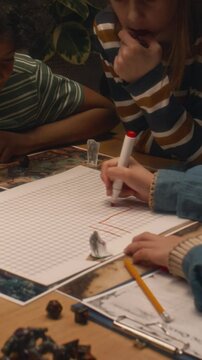 Vertical side shot of kid drawing location map with red marker pen while playing table game with diverse friends at home in evening