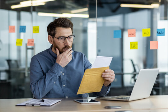 Shocked and worried young businessman man reading received letter in envelope, sitting at desk in office and holding hand on beard