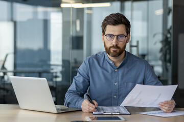 Fototapeta premium Portrait of a young man in glasses and a blue shirt sitting in the office at a table with a laptop, engaged in data analysis and statistics, looking seriously at the camera