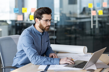 A serious and focused young man, an architect, surveyor and engineer, works in an office with desks...