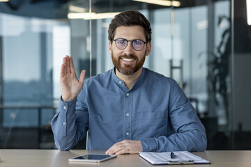 Portrait of a smiling young man in a blue shirt and glasses sitting at a desk in the office, talking on a video call, greeting and waving at the camera