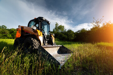 small bulldozer stands in green grass against the backdrop of sun rays in a forest belt at sunset