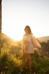 A young woman stands near a rustic stone wall in a lush, outdoor setting at sunset. She is bathed in warm, golden light, creating a serene and contemplative atmosphere.