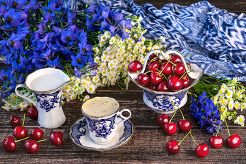 A cup of coffee, a pot for milk, a basket with cherries in blue and white colors, a bouquet of wild flowers and a blue shawl. Summer still life with cherries