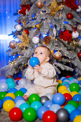 Baby Playing With Balls by Christmas Tree. A baby sits in a ball pit, holding a blue ball in front of a decorated Christmas tree.