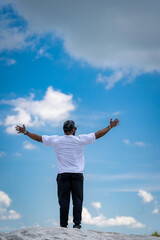 Man Standing On Hilltop With Arms Outstretched. A man in a white t-shirt and black pants stands on a hilltop with his arms outstretched, looking up at the blue sky with white clouds.