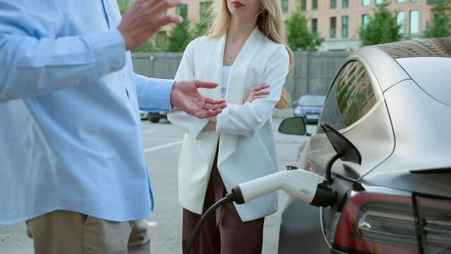 A man and a woman talking near an electric car while it is charging at a charging station in a slow motion
