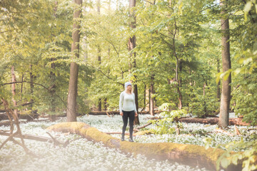 Female traveller with blonde hair and in sportswear walks on a tree trunk surrounded by flowering bear garlic. Hiking lifestyle. Enthralled by the beauty of wild nature