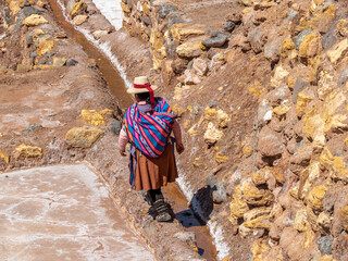 Peruvian woman in traditional colorful dress with hat and bundle on back, walking away from camera through salt mines.