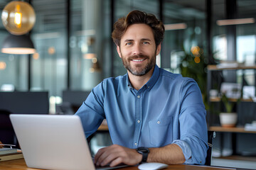 Obraz premium Portrait of a successful and confident young man in a blue shirt sitting in the office at a desk, working on a laptop and smiling at the camera 
