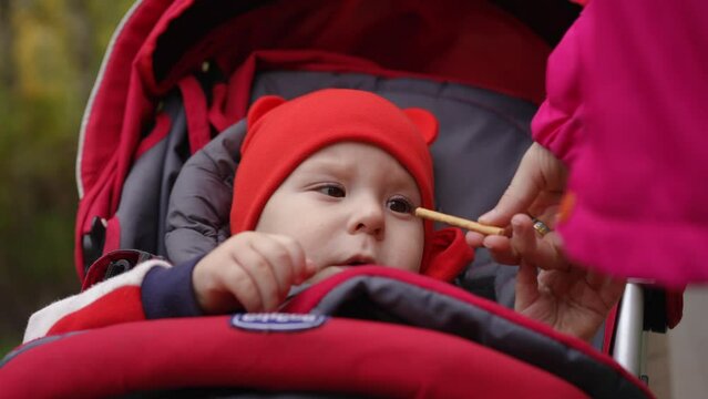 Slow motion. Close-up. A teenage sister gives breadstick to her little brother, who is sitting in a baby carriage. A little boy in a red beanie is eating salt sticks and smiling
