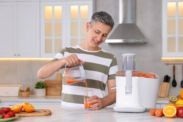 Smiling man pouring juice into glass in kitchen. Juicer and fresh products on white marble table