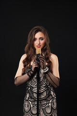 Beautiful young woman in stylish dress with microphone singing on black background