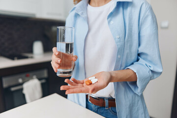 Woman holding glass of water and pill in hand standing in front of kitchen counter, healthcare concept