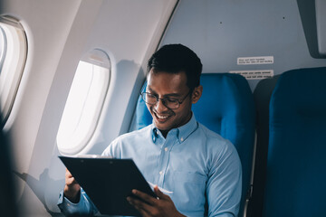 Attractive Asian male passenger of airplane sitting in comfortable seat while working laptop and tablet with mock up area using wireless connection. Travel in style, work with grace.