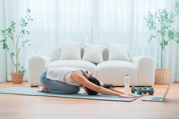 Young Asian woman stretching yoga workout on exercise mat while online training class with computer laptop in living room