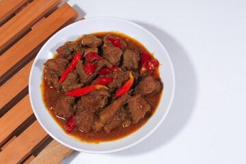 beef cooked in rendang with a garnish of cayenne pepper, served on a white plate and wooden coaster