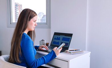 Mujer ejecutiva mirando su teléfono móvil y haciendo trabajo remoto con laptop y agenda sobre escritorio blanco